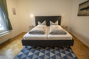 Un lit double avec une tête de lit capitonnée, deux tables de chevet, un tapis à motifs et une fenêtre avec des rideaux.