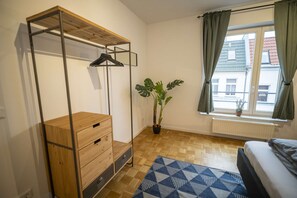 Een kamer met een bed, een houten dressoir, een kledingrek, een potplant en een raam met gordijnen.