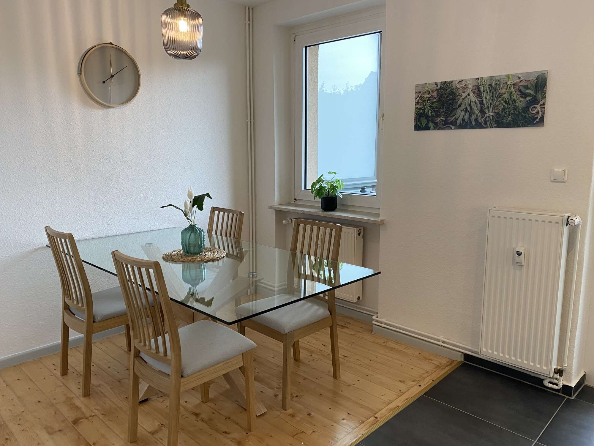 Ett modernt matområde med ett glasbord, trästolar, en vas med blommor, en klocka, en hängande lampa och ett fönster med utsikt.