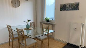 Ett modernt matområde med ett glasbord, trästolar, en vas med blommor, en klocka, en hängande lampa och ett fönster med utsikt.