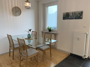 Ett modernt matområde med ett glasbord, trästolar, en vas med blommor, en klocka, en hängande lampa och ett fönster med utsikt.