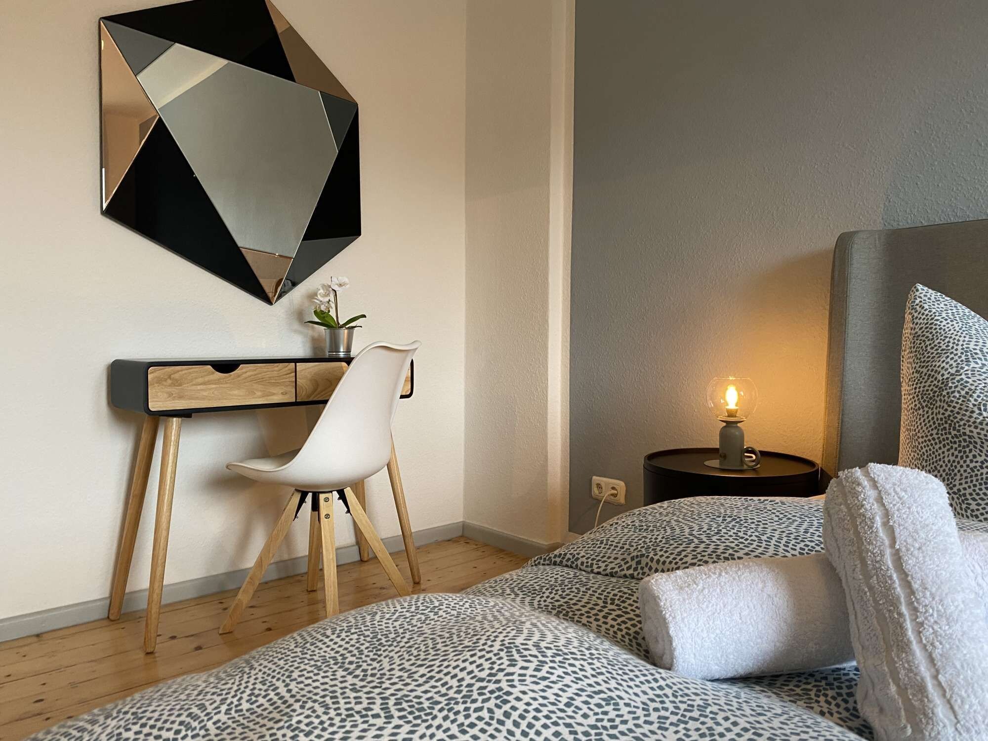 Un dormitorio moderno con una cama, un escritorio, una silla, una mesita de noche con una lámpara y un espejo decorativo.