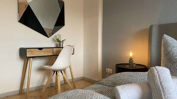 Un dormitorio moderno con una cama, un escritorio, una silla, una mesita de noche con una lámpara y un espejo decorativo.
