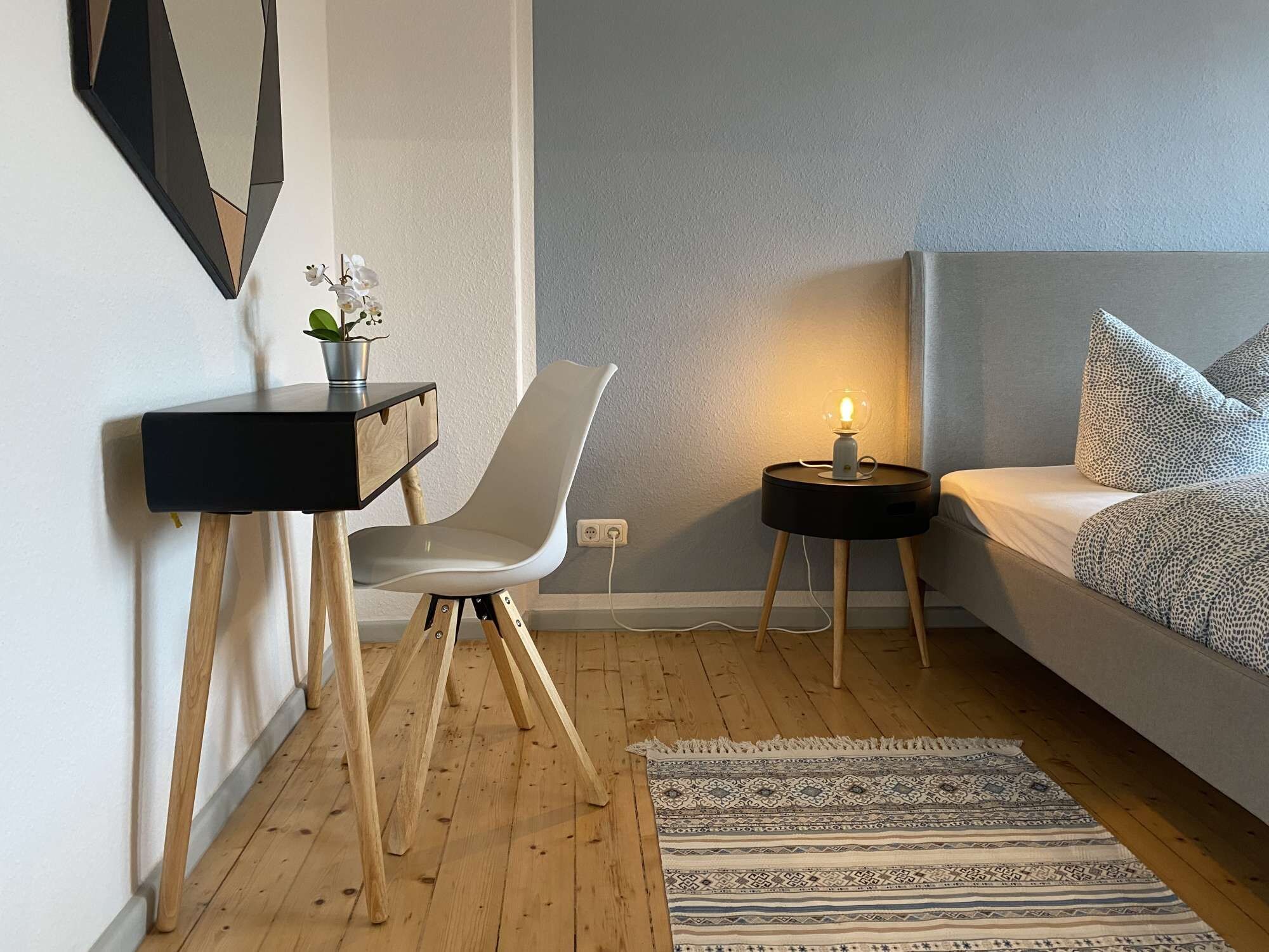 Ett modernt sovrum med en säng, ett skrivbord, en stol, ett litet bord med en lampa och en mönstrad matta.