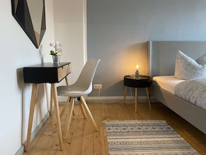 Ett modernt sovrum med en säng, ett skrivbord, en stol, ett litet bord med en lampa och en mönstrad matta.