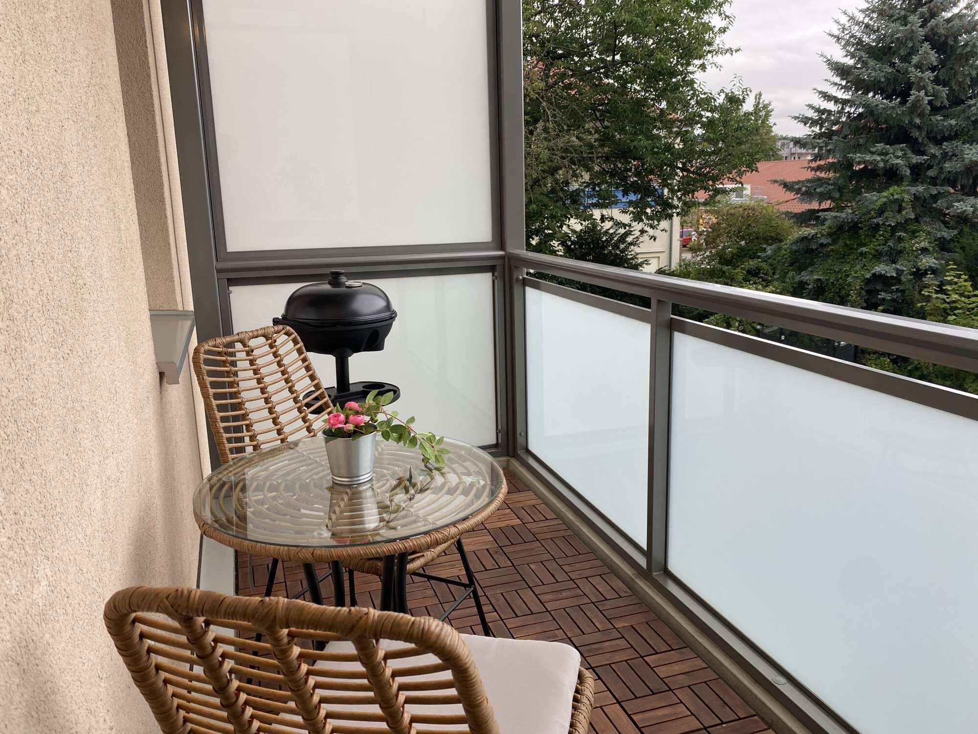 A balcony with wicker chairs, a glass table, and a black barbecue grill.