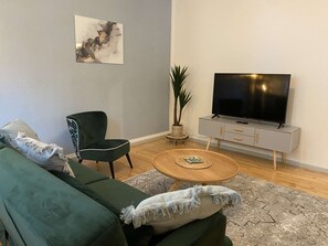 Un saló amb un sofà verd, una cadira, una taula de centre, una televisió sobre un suport i una planta.