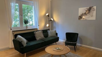 Ett vardagsrum med en mörkgrön soffa, ett runt soffbord i trä och en mörkgrön fåtölj. Det finns ett fönster med vita gardiner och en golvlampa.