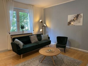 Un saló amb un sofà verd fosc, una taula de centre rodona de fusta i un butaca verda fosca. Hi ha una finestra amb cortines blanques i una làmpada de peu.