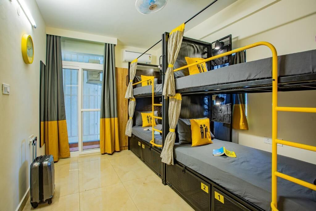 A dormitory room with bunk beds, a suitcase, and a window with curtains.