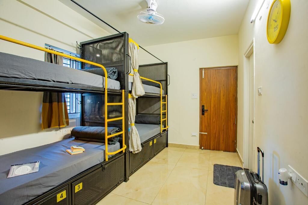 A dormitory room with bunk beds, a wooden door, a suitcase, and a clock on the wall.