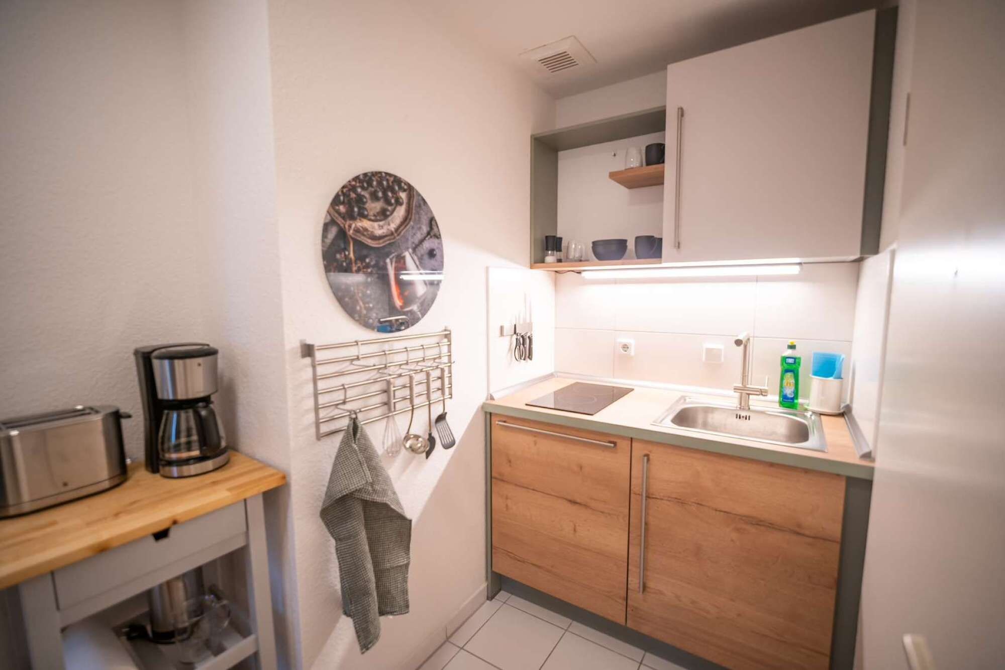 Una cocina compacta con cafetera, un colgador para utensilios, un lavamanos y una encimera con una botella de detergente.
