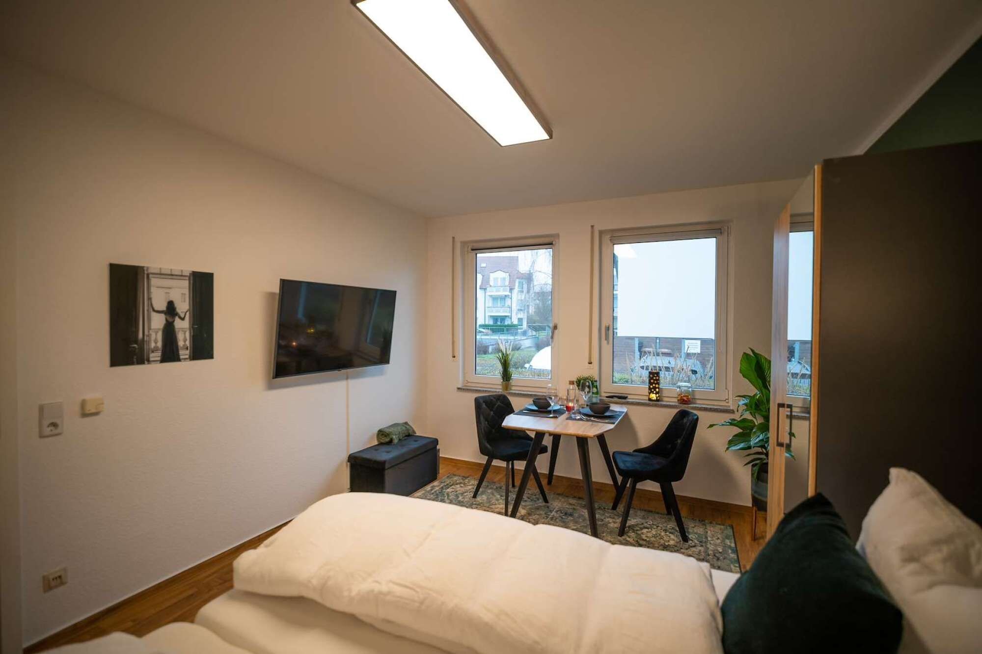 A bedroom with a bed, a desk, two chairs, a TV, and a window with a view of buildings.