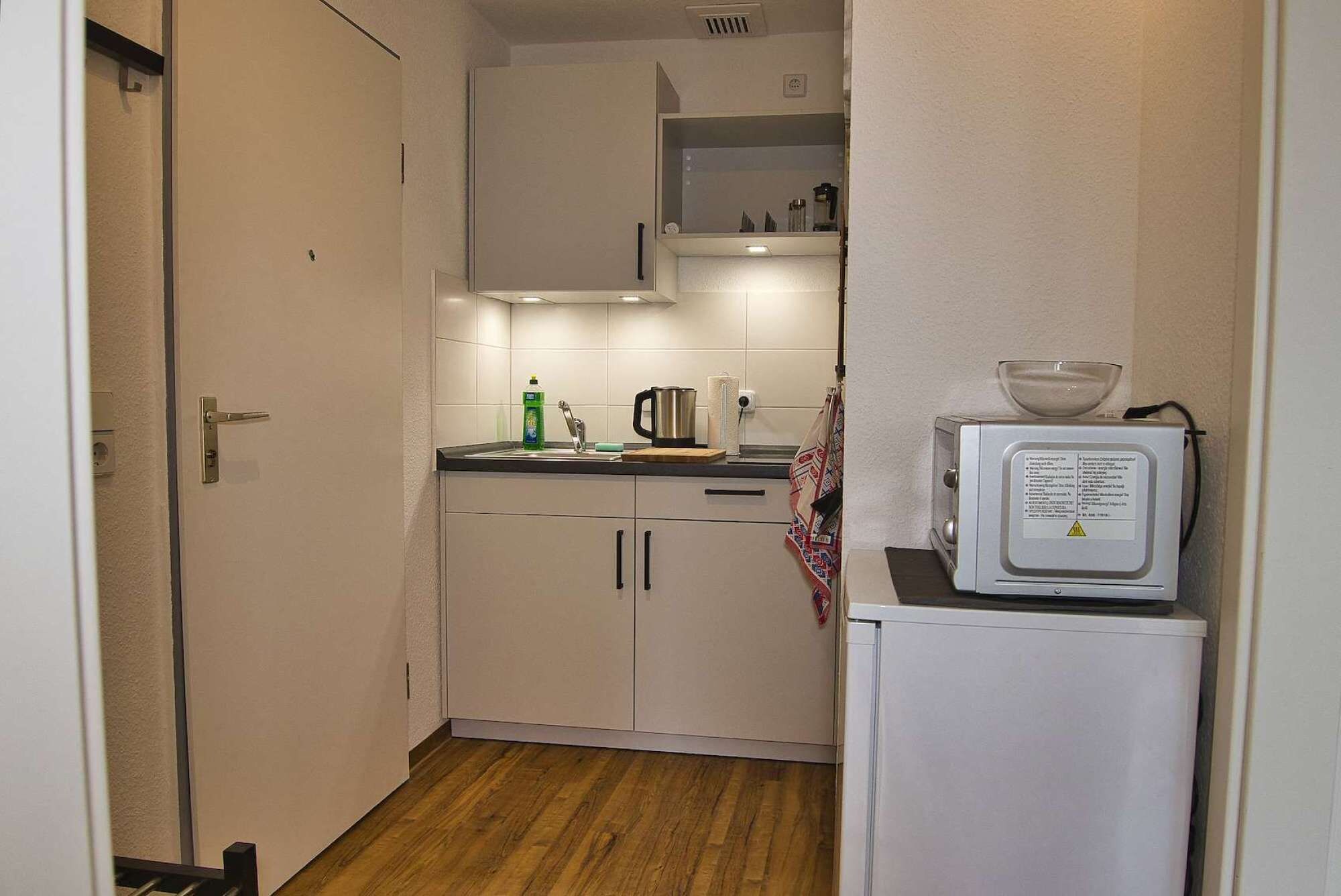 A narrow kitchen with white cabinets, a wooden floor, a kettle, a coffee maker, and a microwave on a small table.