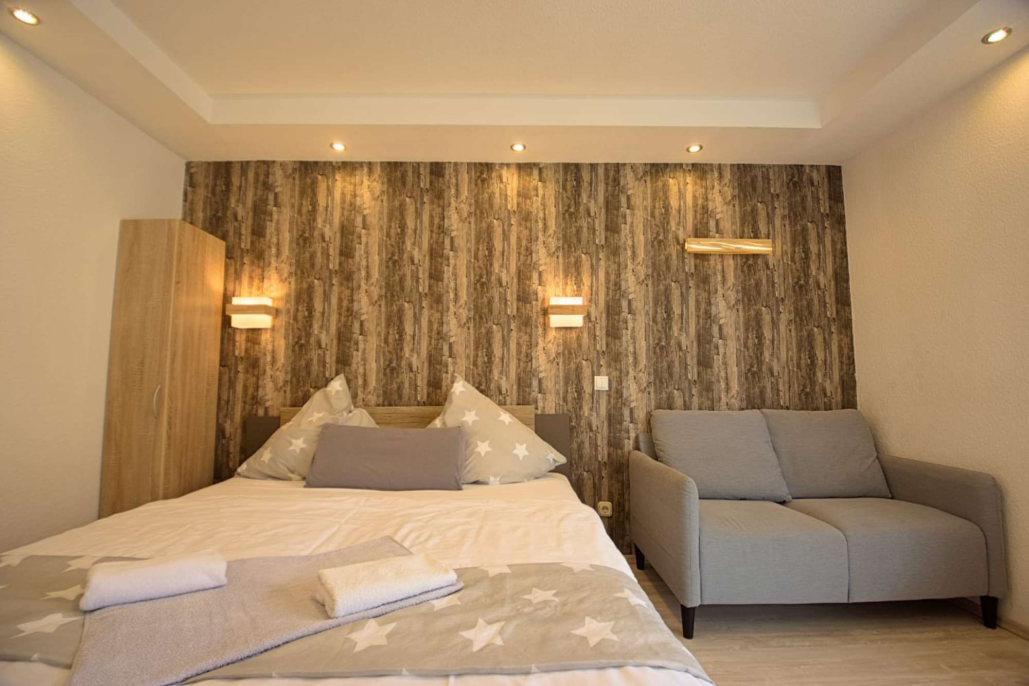 Ein Schlafzimmer mit einem Bett, zwei Nachttischen, einem Sessel und einer Holzwand mit eingelassener Beleuchtung.