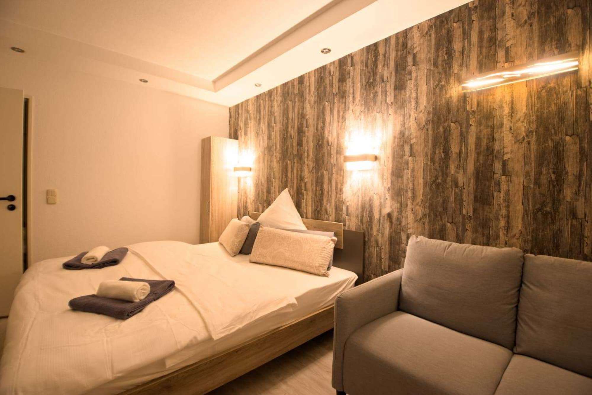 A hotel room with a bed, a sofa, and a wooden accent wall with wall sconces.