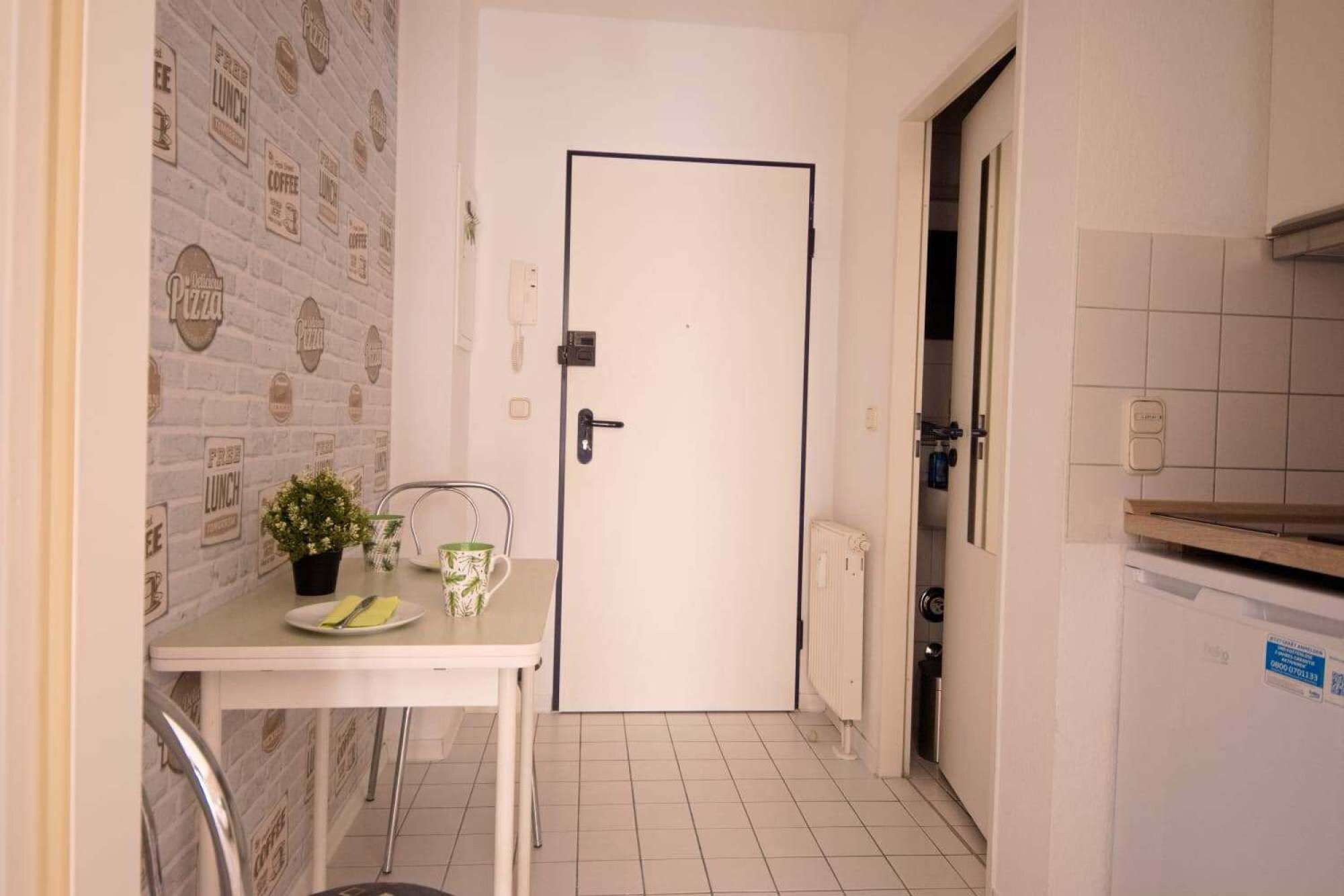 Een smalle gang met een witte deur, een klein tafeltje met een bloempot en een betegelde vloer.