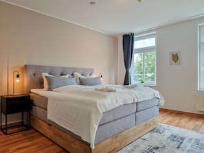 A bedroom with a bed, bedside table, lamp, window, and a view of greenery outside. - RR | Apartments Kappel | 85 m² | Lift | WIFI (Chemnitz)