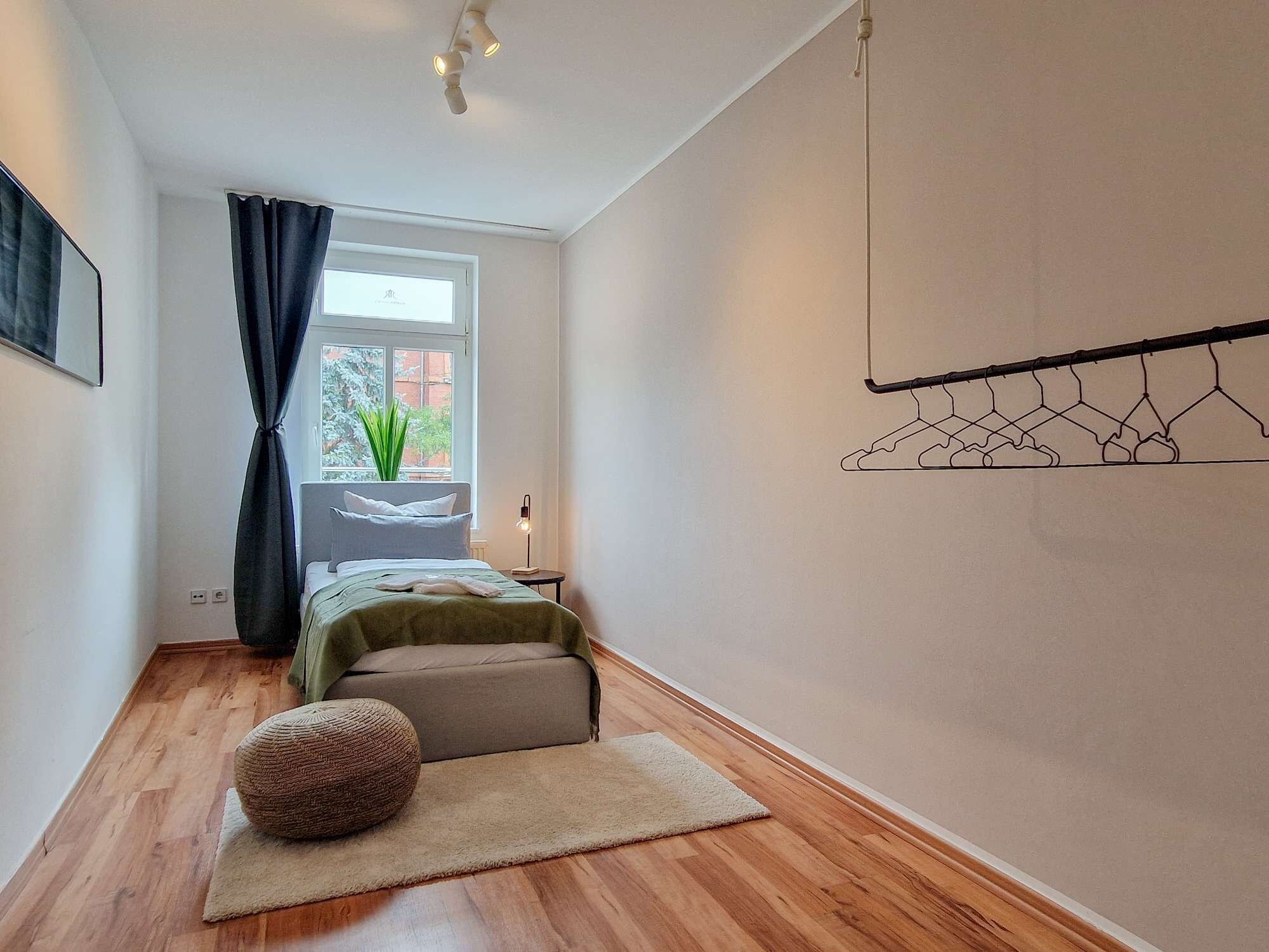 Een slaapkamer met een bed, een nachtkastje, een lamp, een kledingrek en een raam met uitzicht op bomen.