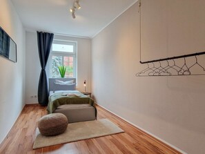A bedroom with a bed, a bedside table, a lamp, a hanging clothes rack, and a window with a view of trees.