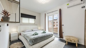A modern bedroom with a bed, bedside tables, a small stool, a potted plant, and a view of the outside. - Modern Apartment With Scenic Views Earthy Vibes (Shkodër)