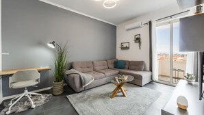 A modern living room with a grey sofa, a glass coffee table, a desk with a chair, and a large window offering a city view. - Modern Apartment With Scenic Views Earthy Vibes (Shkodër)