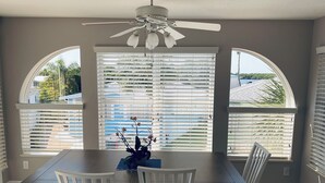 A dining area with a table and chairs, a ceiling fan, and large windows with blinds.