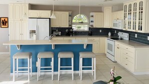 A modern kitchen with a large island, bar stools, white cabinetry, and a view of greenery outside.