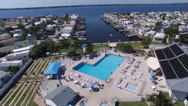 An aerial view of a resort with a central swimming pool surrounded by lounge chairs and umbrellas, a marina with boats, and residential buildings in the background.