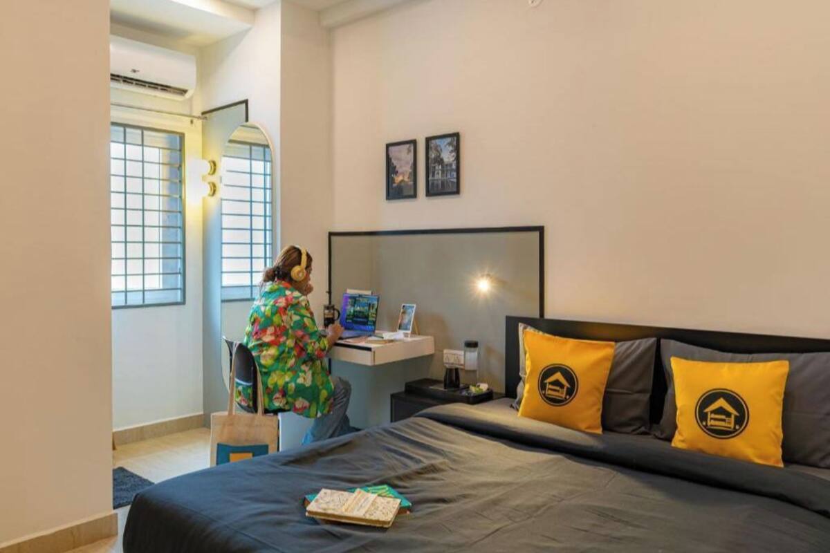 Une chambre d’hôtel avec un lit, un bureau et une chaise. Il y a une fenêtre avec des stores, un miroir fixé au mur et deux tableaux encadrés.