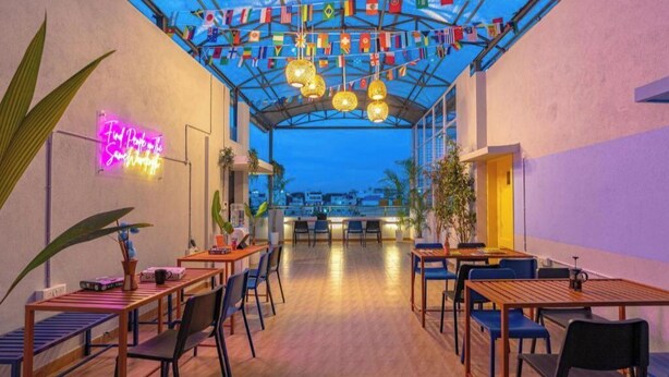 A restaurant with wooden tables and chairs, a neon sign, and flags hanging from the ceiling. - The Hosteller Bangalore, Koramangala (Karnataka)
