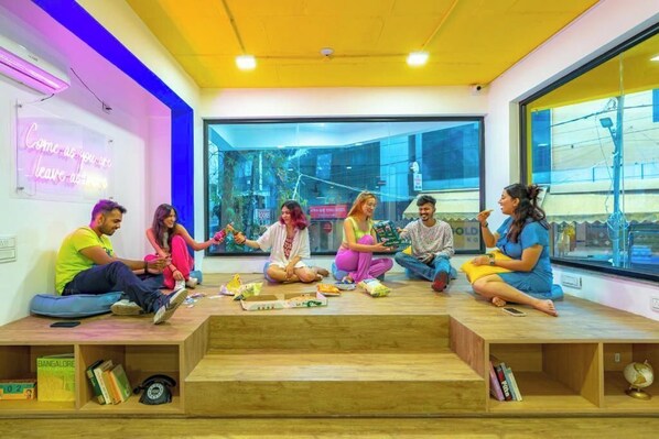 A group of people sitting on wooden steps in a modern room with large windows and a neon sign. - The Hosteller Bangalore, Koramangala (Karnataka)