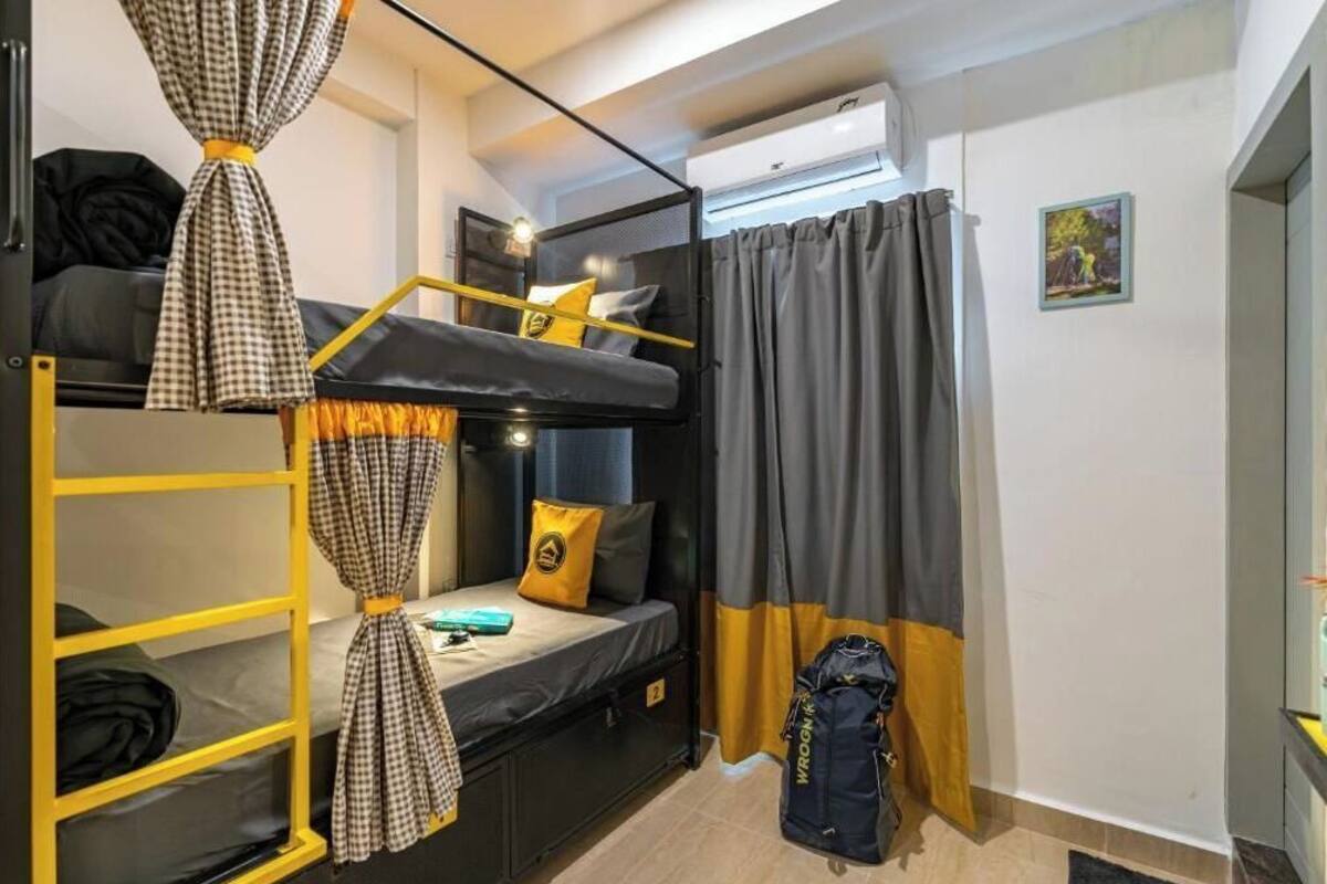 Une petite pièce avec un lit superposé, un bureau et une chaise. Une valise est posée sur le sol et un appareil de climatisation est fixé au mur.