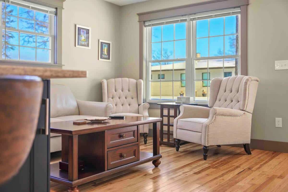 A room with a wooden coffee table, two armchairs, and a sofa. There are two windows with a view of a house outside.