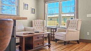 Una habitación con una mesa de centro de madera, dos sillones y un sofá. Se ven dos ventanas con vista a una casa.