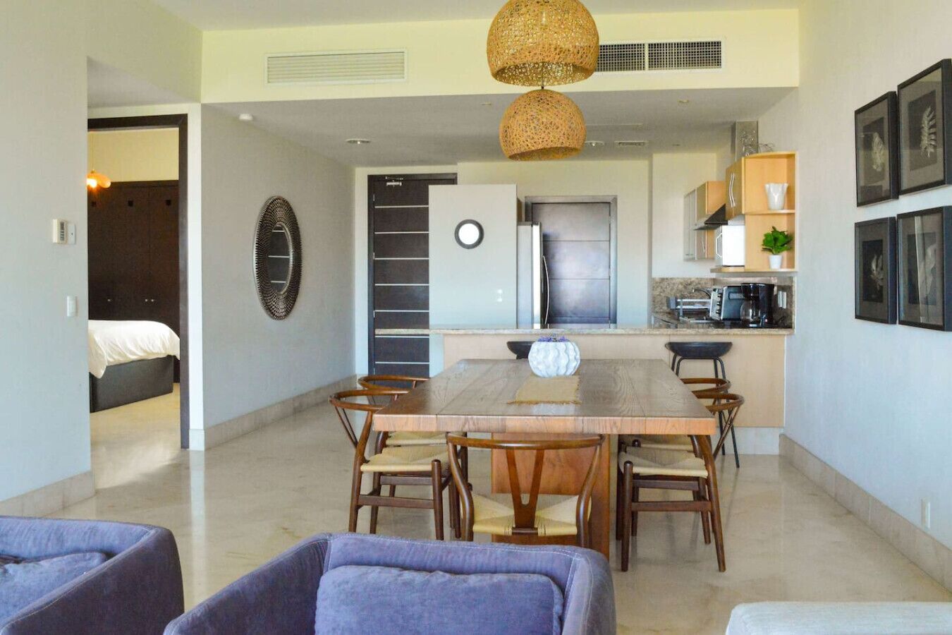 A modern kitchen with a central island, a dining area with a wooden table and chairs, and a visible bed in the adjacent room.
