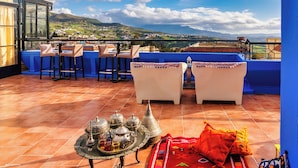 A terrace with a view of a town and mountains, featuring a table with traditional items and a red patterned rug.