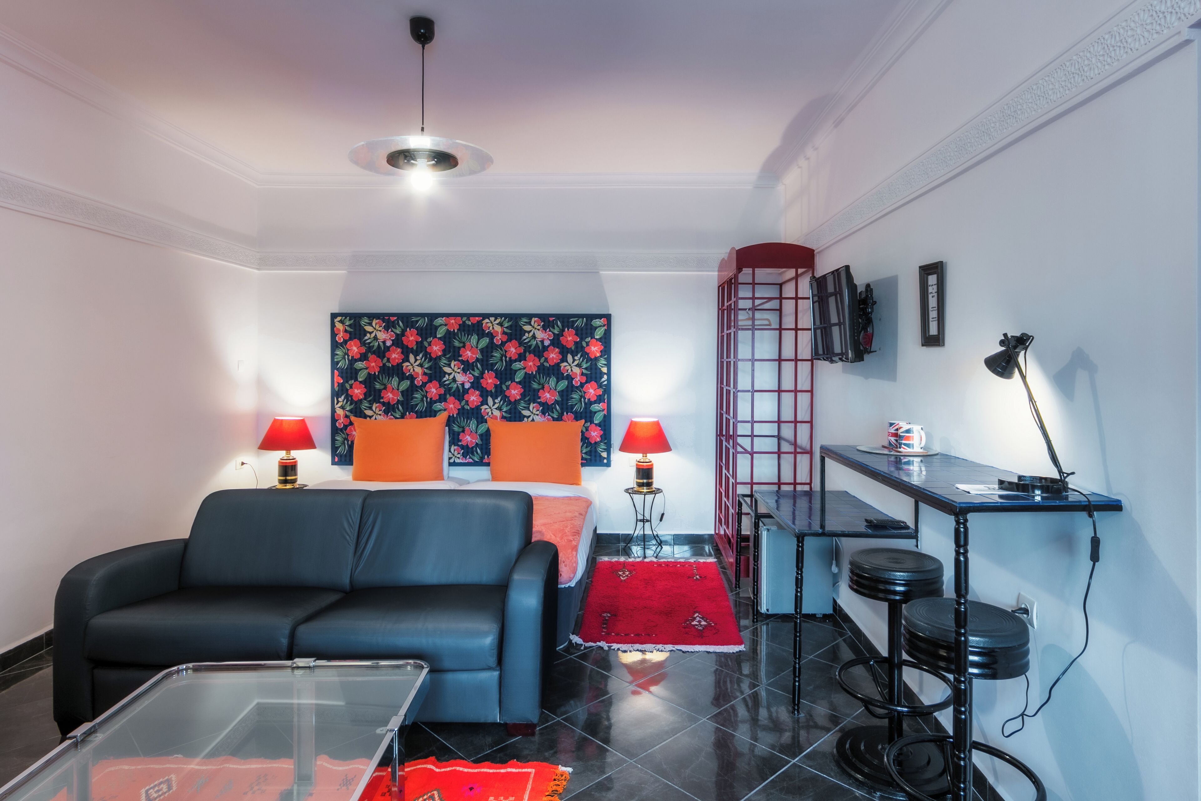 Una sala moderna con un sofá negro, una mesa de centro de vidrio, una alfombra roja, una cama con almohadas naranjas y una escalera roja apoyada en la pared.