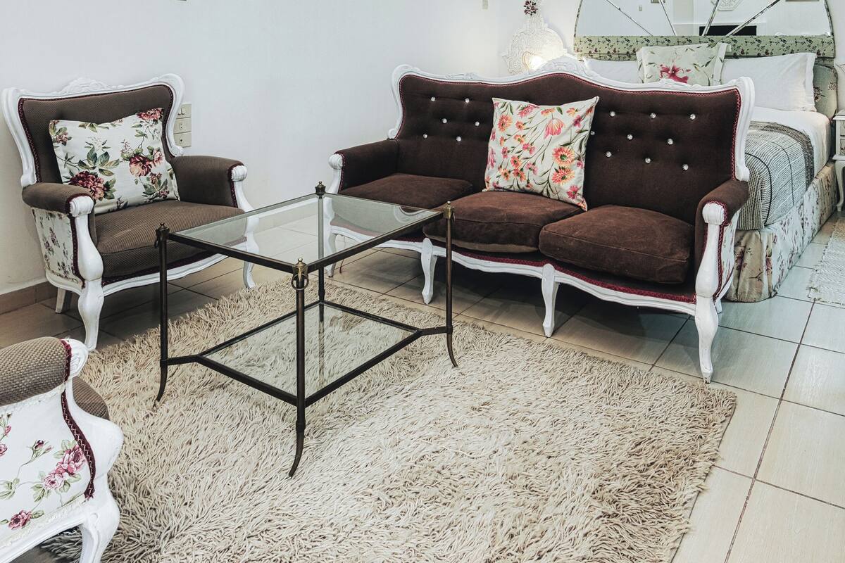 A living room with a sofa, armchairs, a glass coffee table, a patterned rug, a chandelier, and a mirror on the wall.