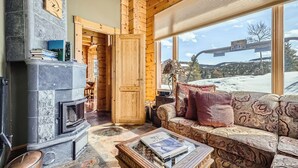 L’intérieur d’une cabane en rondins, avec une cheminée en pierre, un canapé agrémenté de coussins à motifs et une vue sur un paysage enneigé, visible à travers de grandes fenêtres.