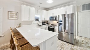 A modern kitchen with white cabinetry, a large island, stainless steel appliances, and a dining area with wicker chairs. - Community Pools | LSV | Close to Beach (Santa Rosa Beach)