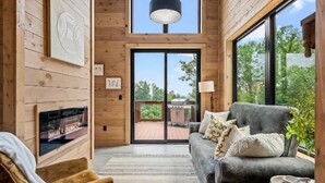 A cozy living room with a fireplace, a sofa, and a large window offering a view of the outdoors. - Stunning Cabin for 6 with Hot Tub & Pool Table! (Sevierville)