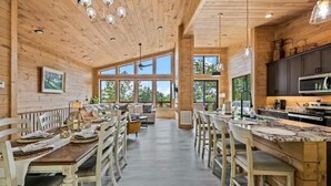 A spacious dining area with wooden tables and chairs, a kitchen with dark cabinets and a large island, and large windows allowing natural light. - Stunning Cabin for 6 with Hot Tub & Pool Table! (Sevierville)