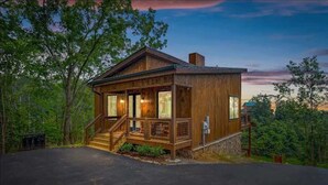 A wooden cabin with a porch, surrounded by trees. - Stunning Cabin for 6 with Hot Tub & Pool Table! (Sevierville)