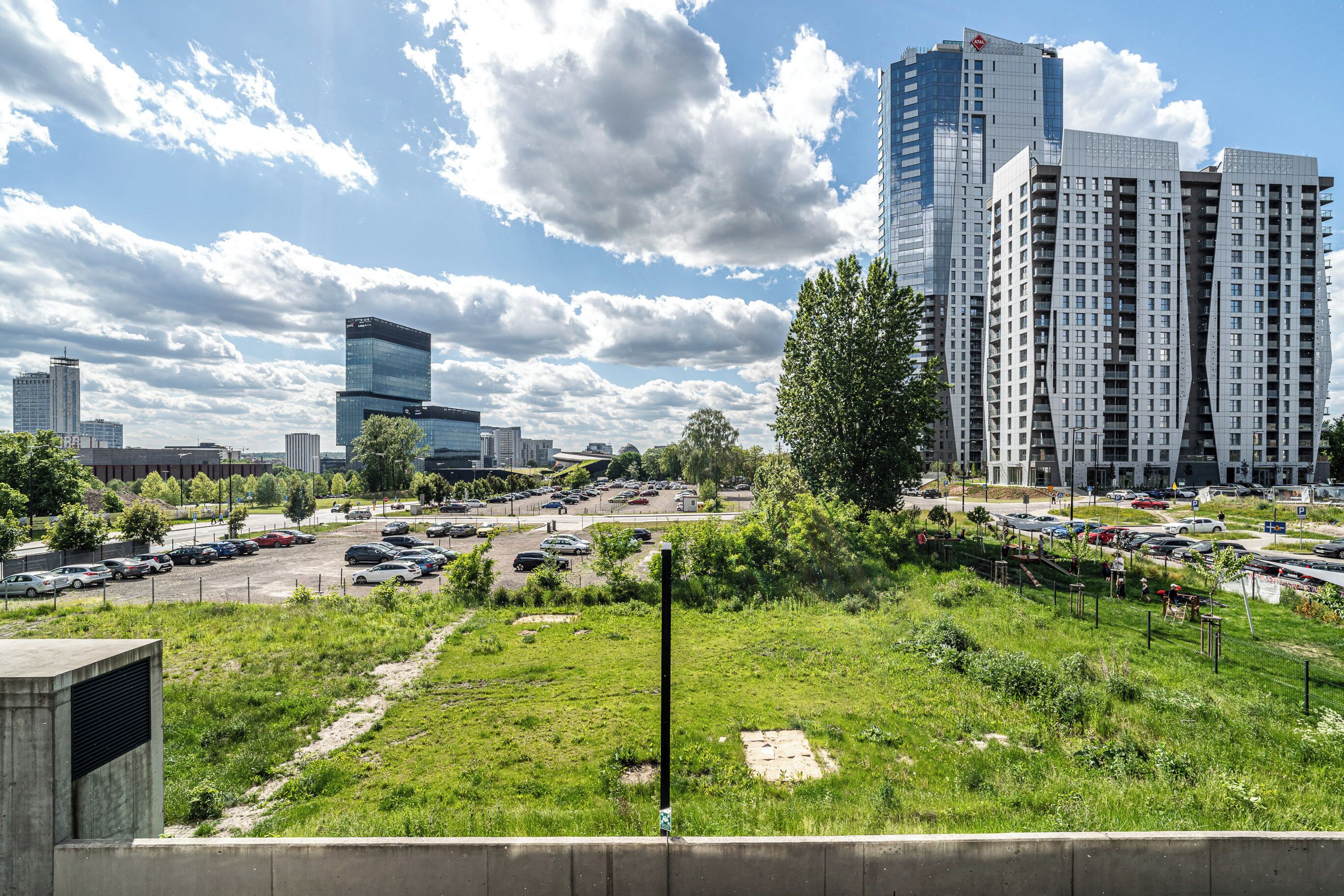 A cityscape with modern high-rise buildings, a parking lot, and a grassy area.
