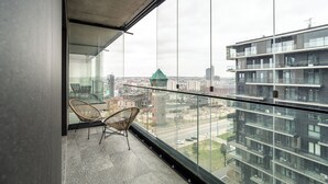 Un balcon situé dans un immeuble de grande hauteur, doté d'une balustrade en verre, de fauteuils en osier et offrant une vue sur le paysage urbain.