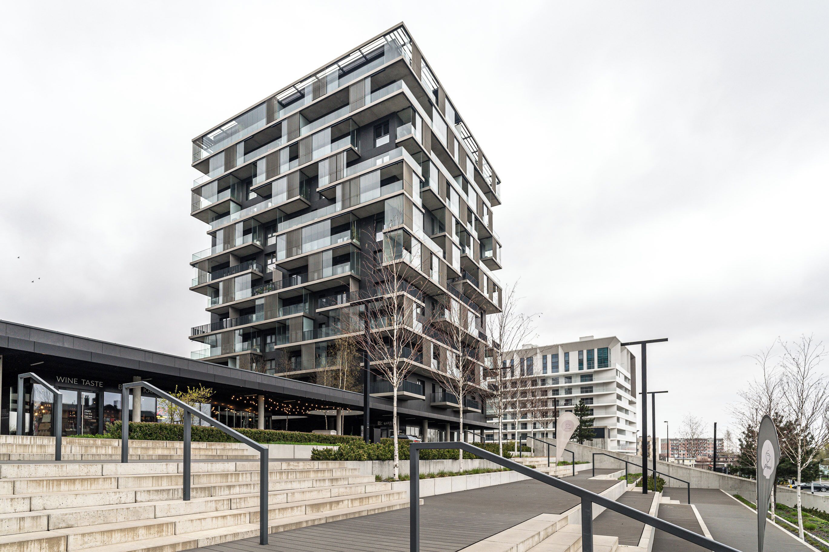 A modern multi-story building with balconies, a covered entrance, and a landscaped area with steps and railings.