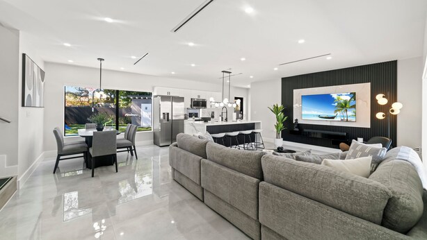 A modern living room with a large grey sectional sofa, a dining table with chairs, a kitchen area, and a TV mounted on the wall. - Modern 5BR Dream • Courts, Playground • Pool_BBQ (Miami)