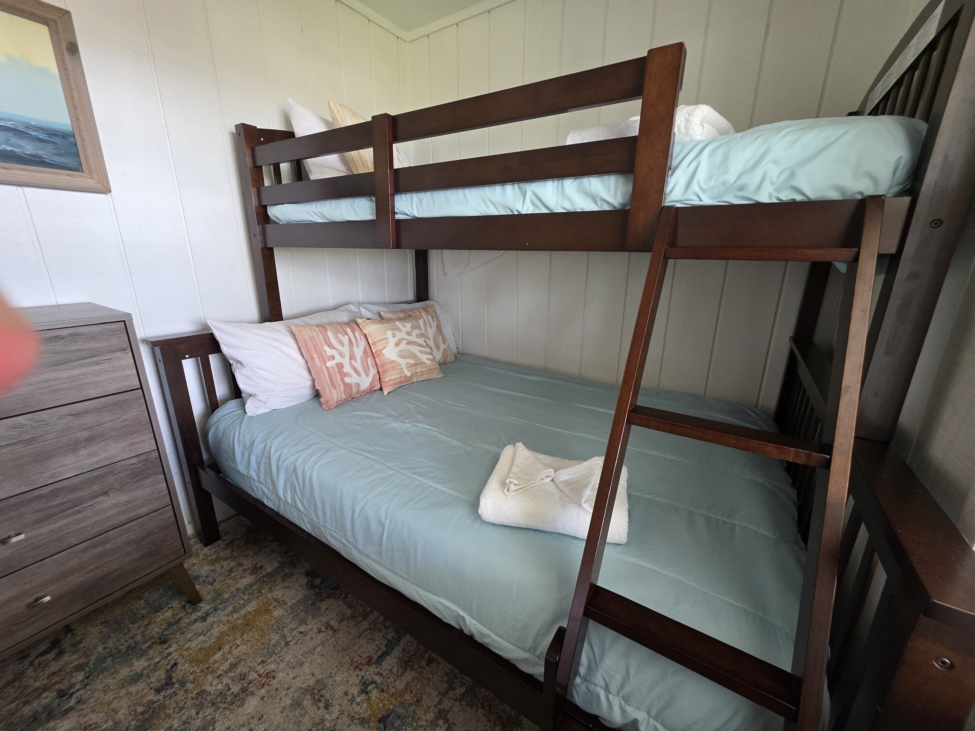 A bunk bed with a blue mattress and white pillows, a wooden dresser, and a framed picture on the wall.