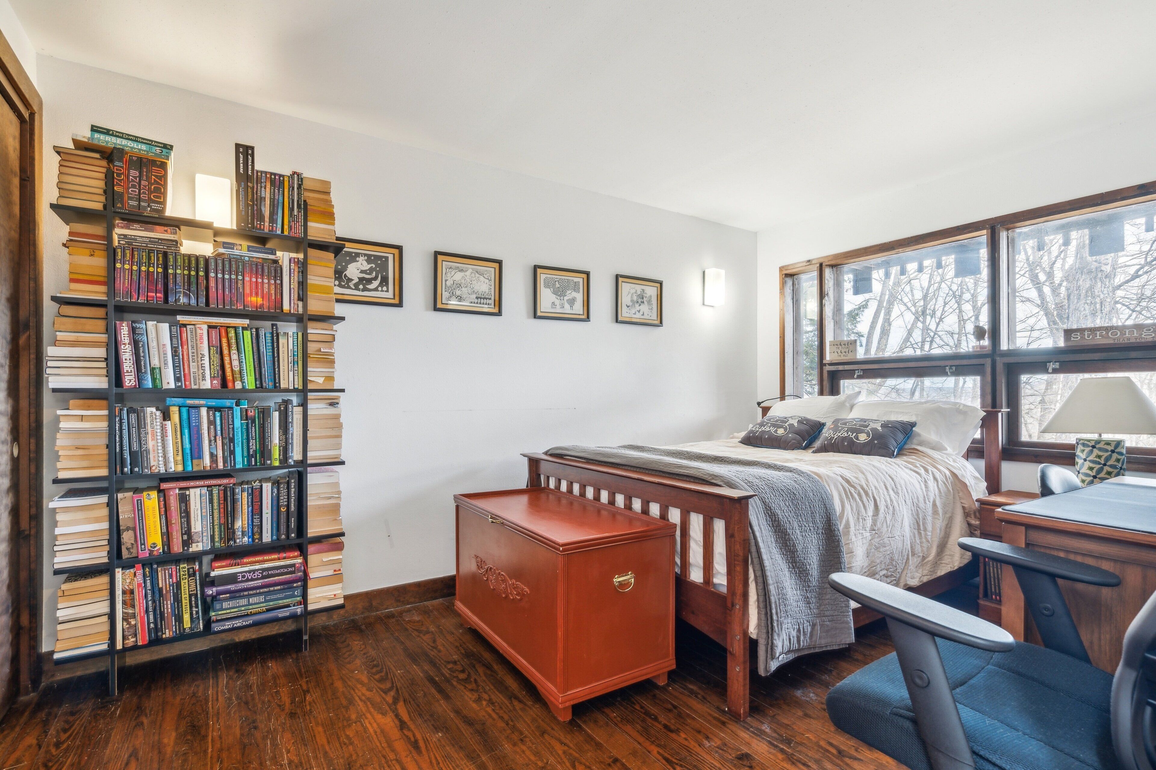 A bedroom with a large bookshelf, a bed with a wooden headboard, a desk with a chair, and a window with a view of trees.
